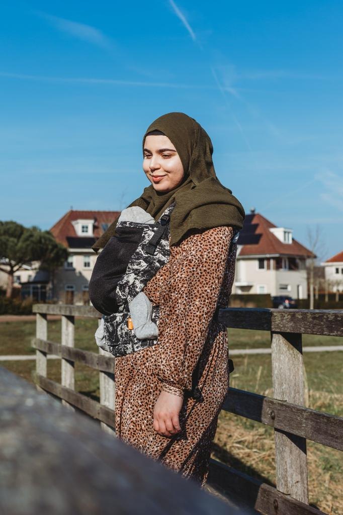 Feride staat bij een houten hek en glimlacht, met haar baby in een draagzak