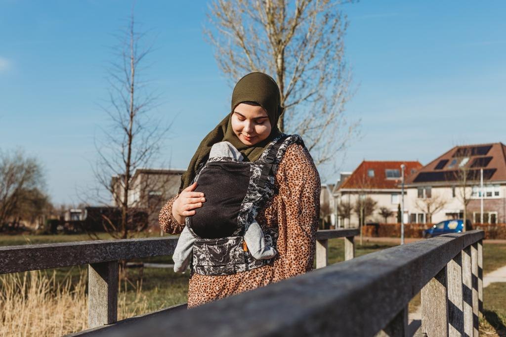 Feride bij een houten hek, ze kijkt naar haar baby in de draagzak