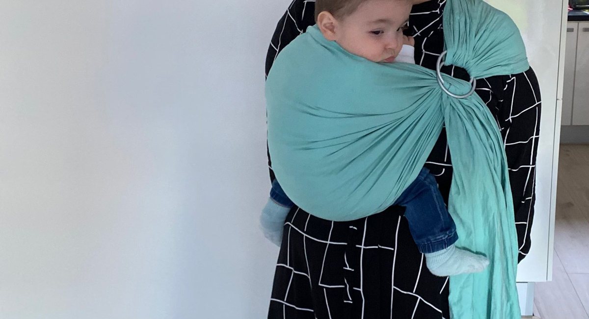 Een kind in een turquoise ringsling binnenshuis, het kijkt opzij